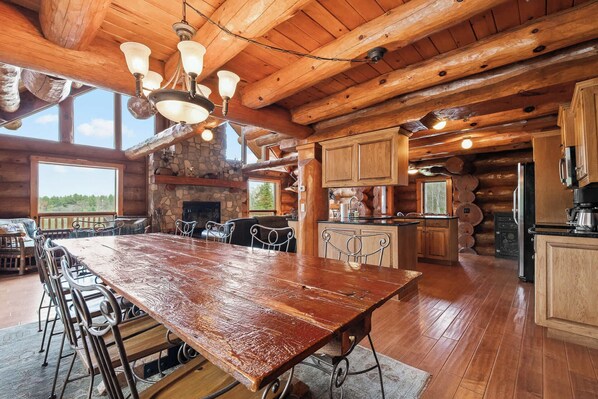 A spacious log cabin interior with a large wooden dining table, stone fireplace, and a kitchen area.