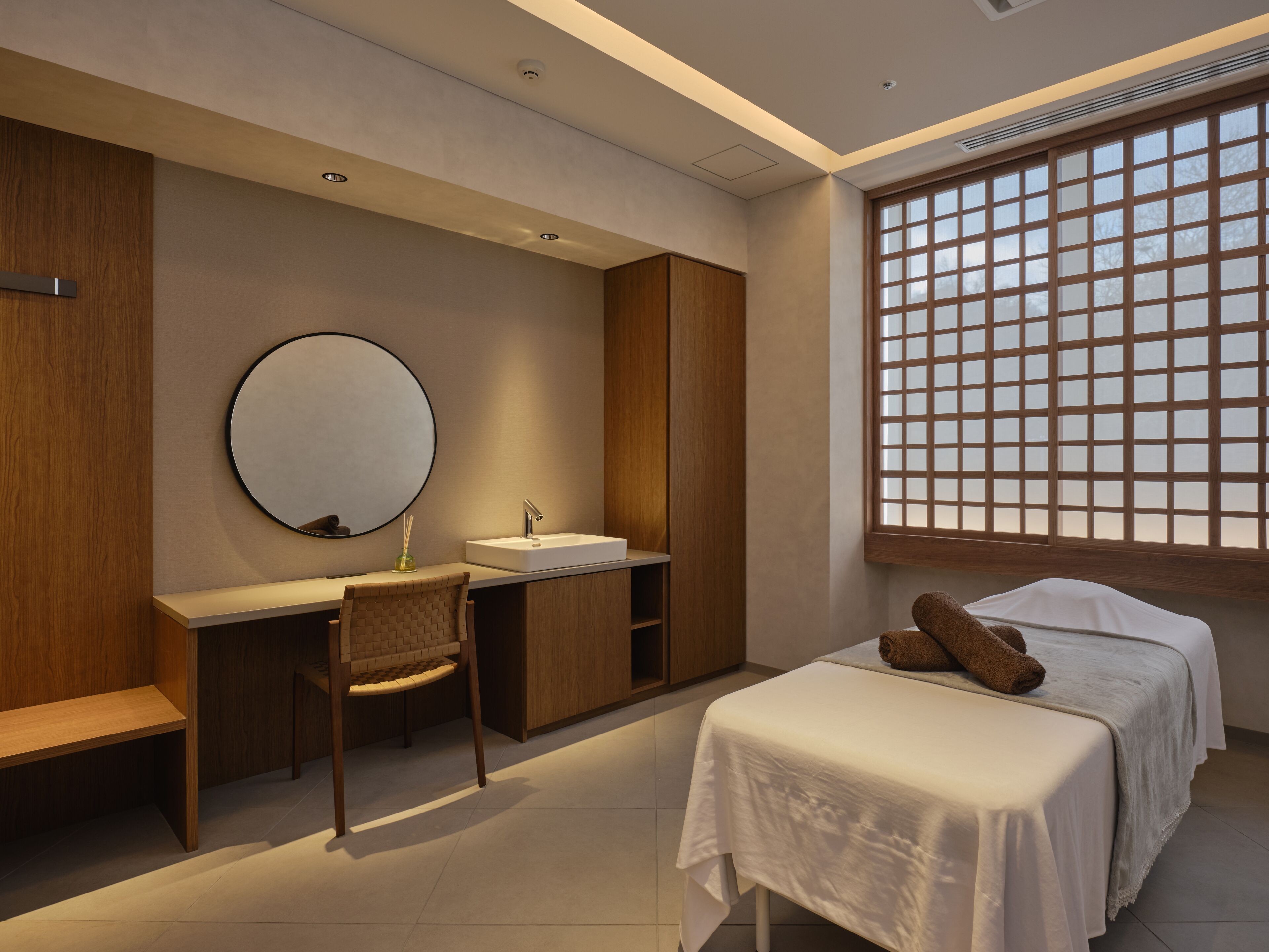 A modern bedroom with a desk, a round mirror, a bed with a white bedspread, and a window with a wooden grid.
