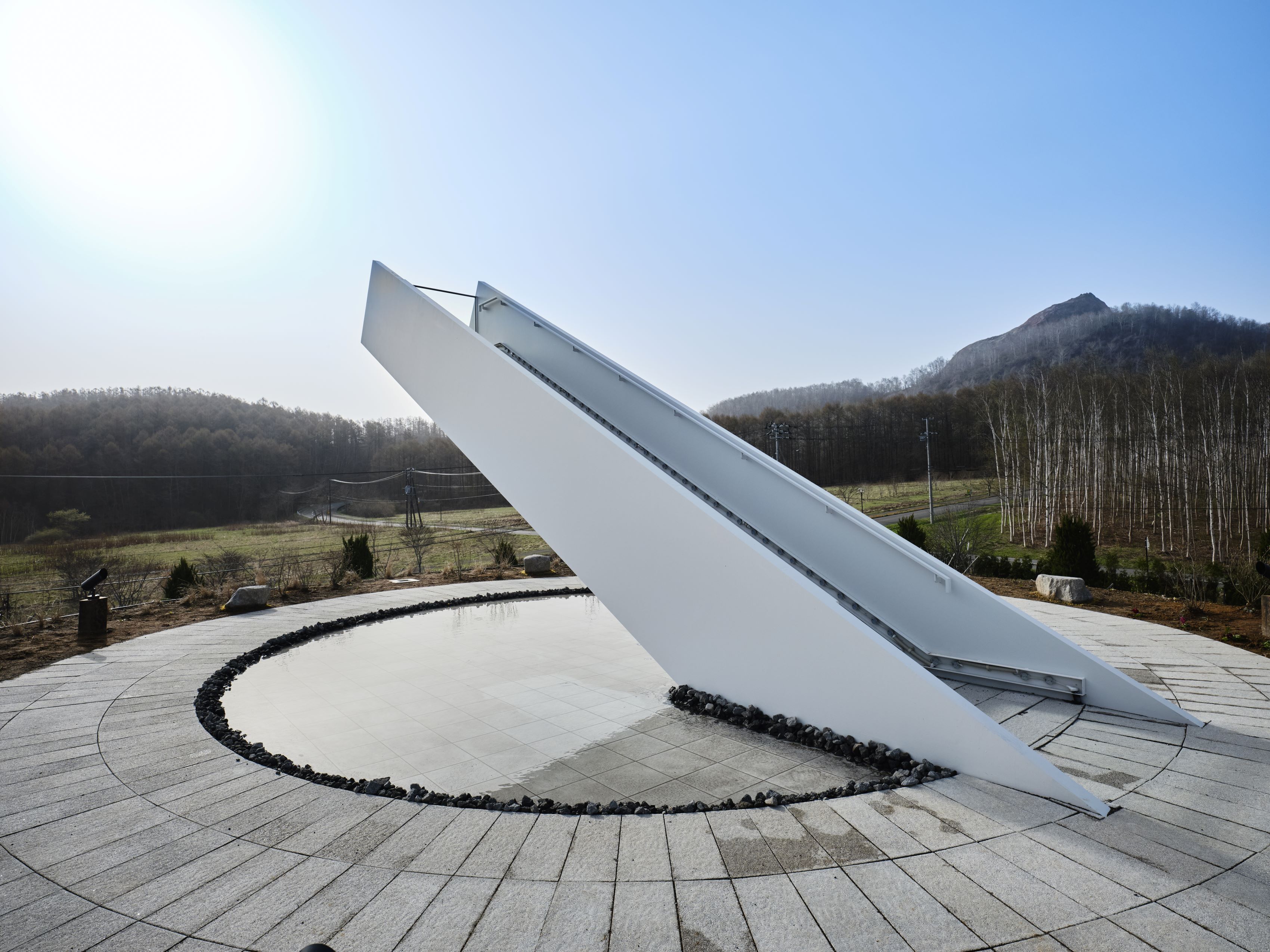 Una estructura arquitectónica moderna con un panel blanco inclinado, rodeada por un sendero circular de piedra y una fuente de agua reflectante, frente a un cerro cubierto de bosque y un cielo despejado.
