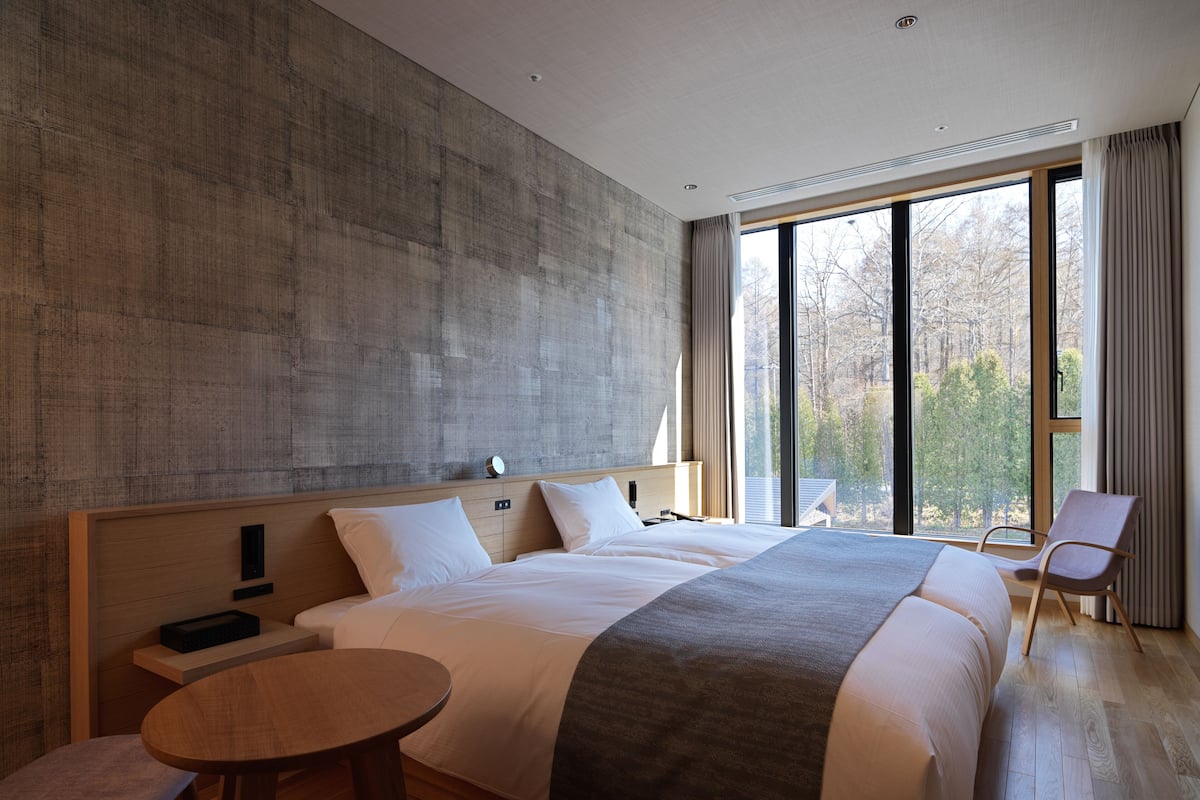 Habitación de hotel moderna con una cama grande, una mesita redonda y vistas a árboles a través de amplias ventanas.