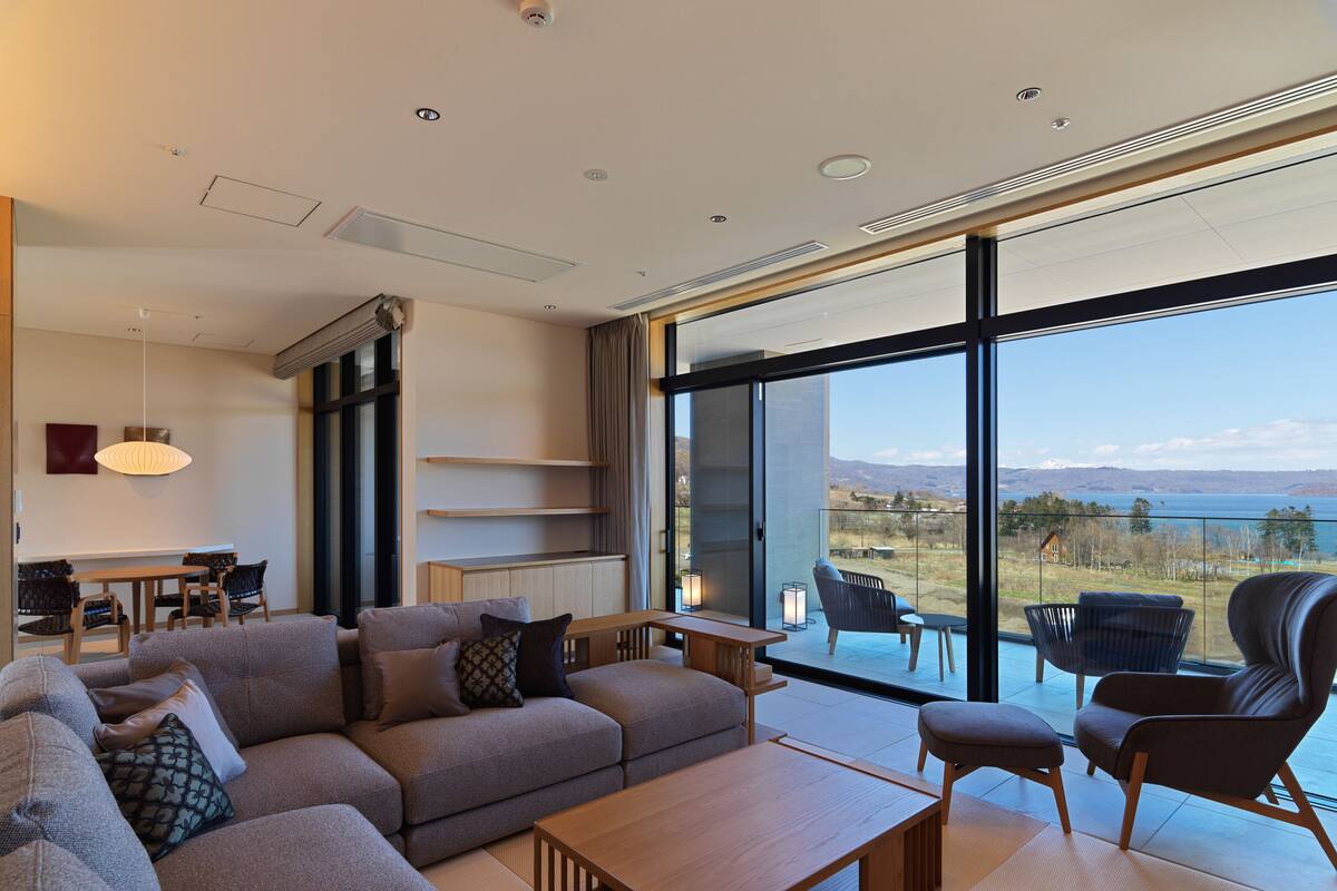 Una sala de estar moderna con sofá, mesa de centro y sillas, ventanas amplias con vistas a un lago y montañas.