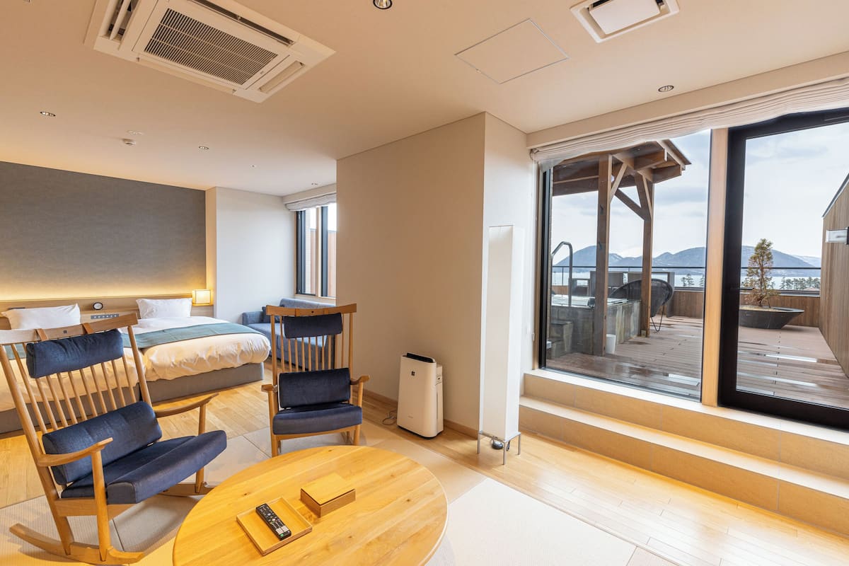 Habitación de hotel moderna con una cama grande, zona de estar con sillas, una mesita y un ventanal con vistas a un balcón y montañas.