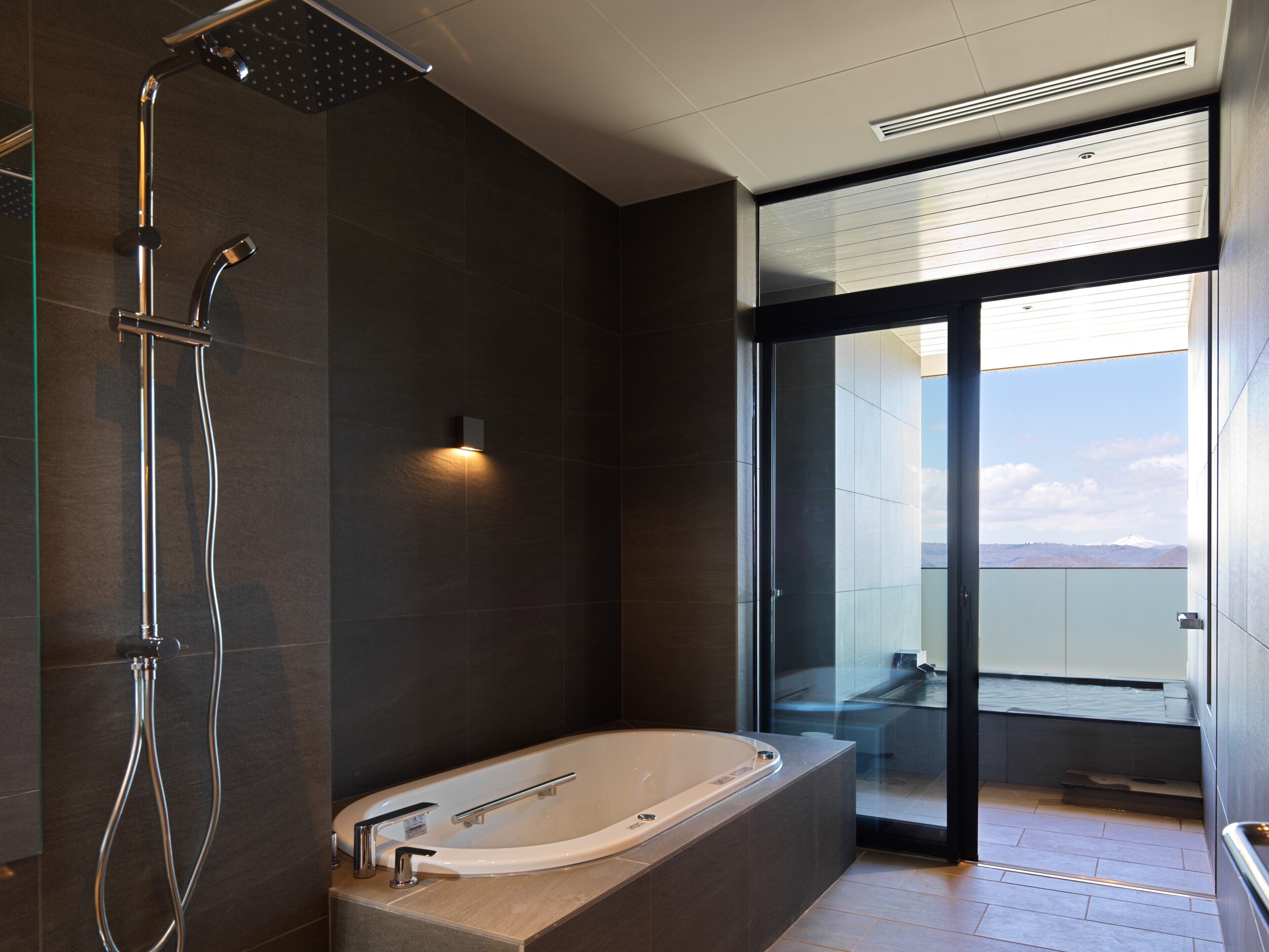 A modern bathroom with a large bathtub, a showerhead, and a glass door leading to a balcony with a view of a mountain.