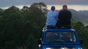 A blue Suzuki vehicle with two passengers on a road surrounded by lush greenery, overlooking a body of water and distant hills during sunset. - a very interesting tour held on top of Mount Batur, 4wd jeeps tour (Bali)