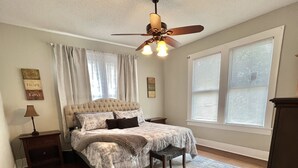 A bedroom with a bed, a ceiling fan, a nightstand, a lamp, and a window with blinds. - Historical Spacious 1-Bedroom Apartment Unit 3 (Shreveport)