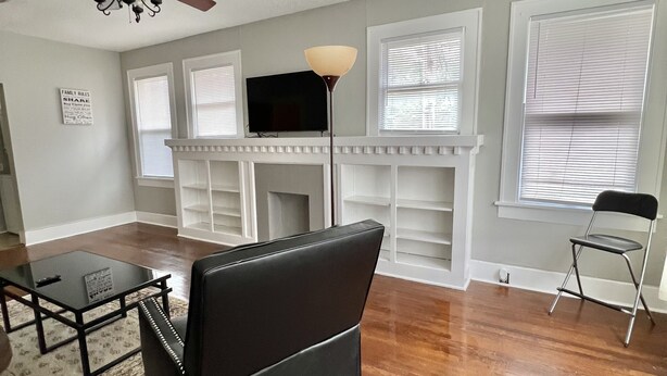A living room with hardwood flooring, a black sofa, a glass coffee table, a fireplace, and a ceiling fan. - Historical Spacious 1-Bedroom Apartment Unit 3 (Shreveport)