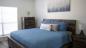 A bedroom with a large bed, a nightstand with a lamp, a wooden dresser, and a painting on the wall. - Safe, Central, Cozy Home with Private Parking, KING Bed, near DFW Airport & more (Euless)