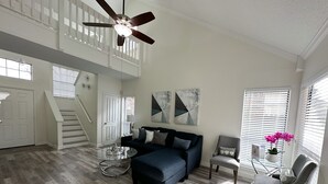 A spacious living room with a dark blue sofa, a glass coffee table, and a grey armchair. A ceiling fan is visible, and there are abstract wall art pieces. - Safe, Central, Cozy Home with Private Parking, KING Bed, near DFW Airport & more (Euless)