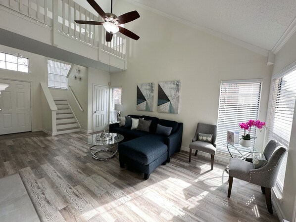 A spacious living room with a dark blue sofa, a glass coffee table, and a grey armchair. A ceiling fan is visible, and there are abstract wall art pieces.