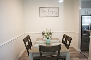 A dining area with a wooden table, four chairs, and a vase with flowers. A sign on the wall reads "Our happy PLACE".