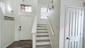 A clean, modern hallway with a wooden staircase, white walls, and a small window with blinds. - Safe, Central, Cozy Home with Private Parking, KING Bed, near DFW Airport & more (Euless)