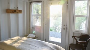 A room with a bed, a wooden chair, a coat rack, and a window with a view of greenery. - Cabin 3 | Rural Oasis on Pond with Panoramic Views (Latah County)