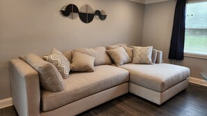 Un canapé d’angle beige agrémenté de coussins à motifs, une œuvre d’art abstraite accrochée au mur et une fenêtre ornée de rideaux foncés.