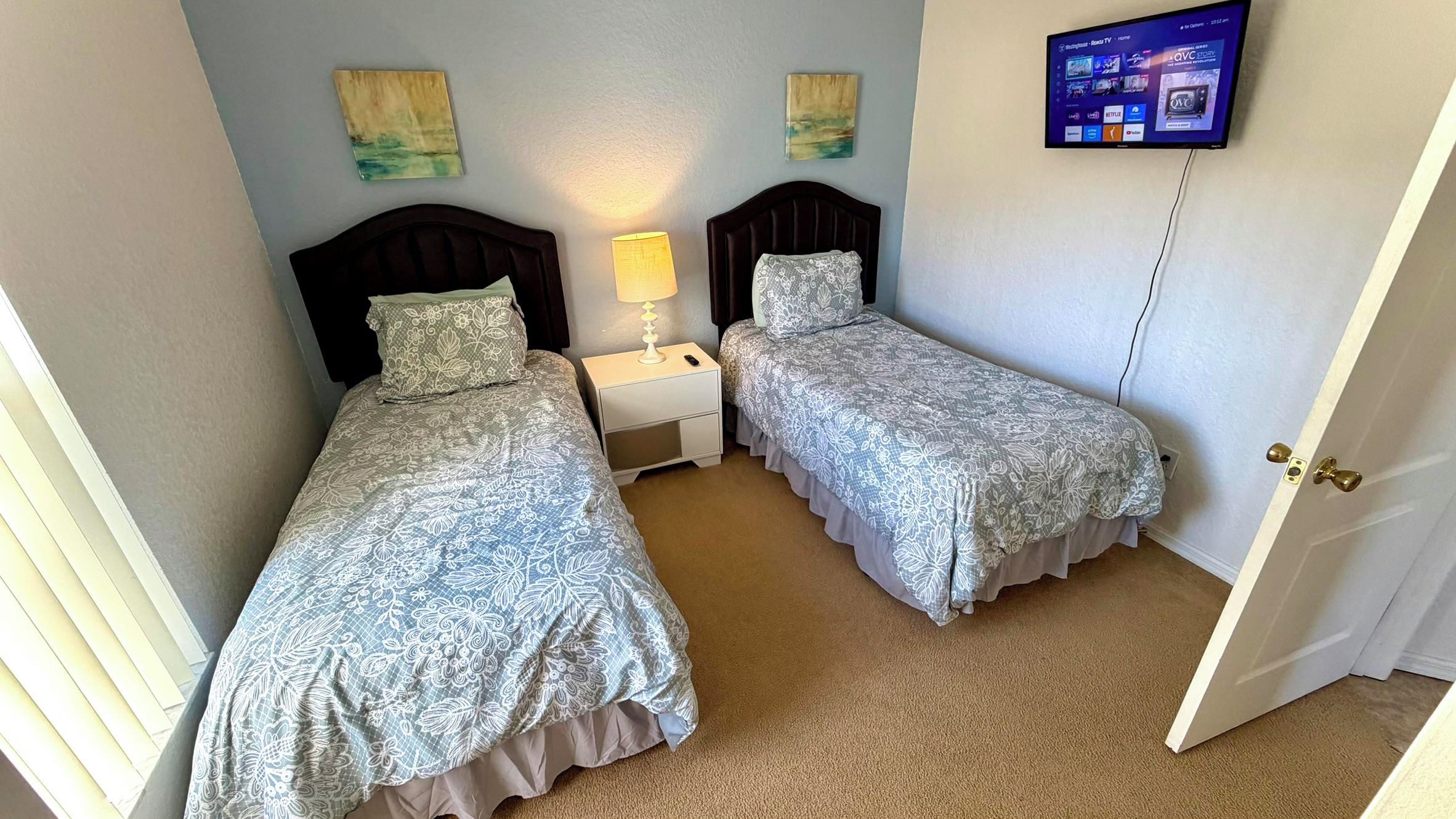 Une chambre d’hôtel avec deux lits simples, une table de chevet avec une lampe, un téléviseur fixé au mur et des œuvres d’art accrochées aux murs.