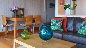 Ett vardagsrum med ett soffbord i trä, en soffa med färgglada kuddar, en matplats med stolar och en vägg med ett dekorativt bladmotiv.
