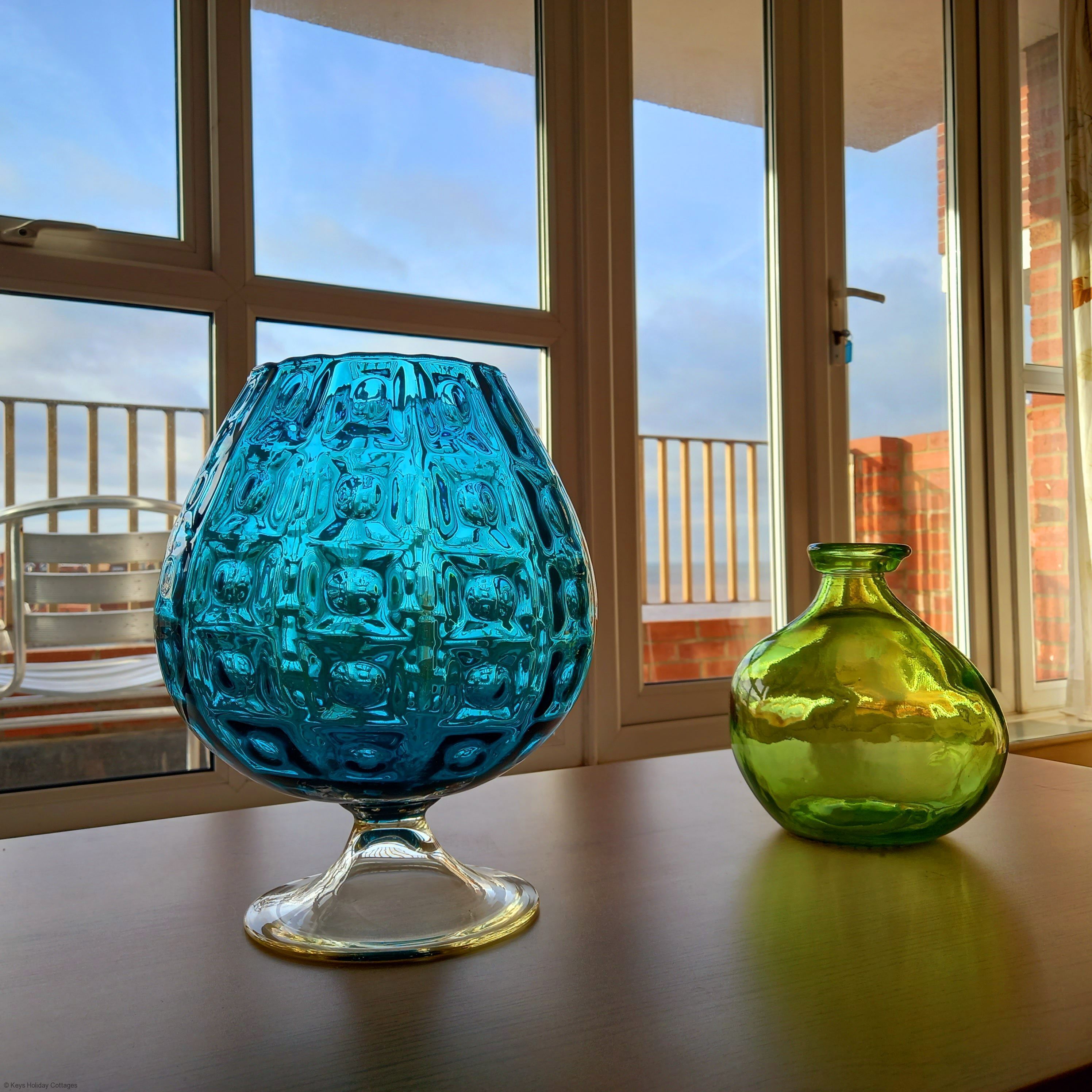 A blue and a green glass vase on a table by a window with a balcony view.