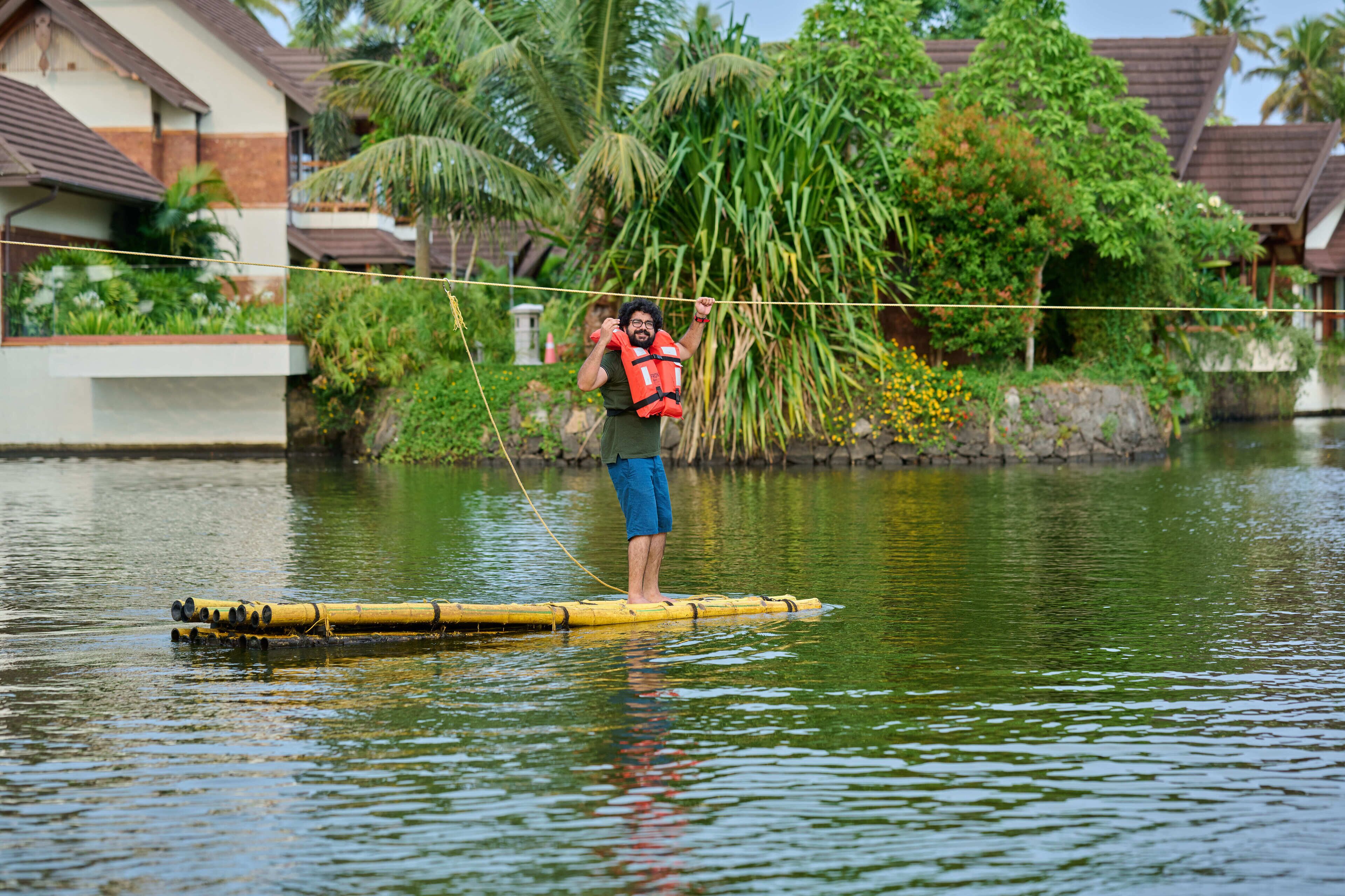 Una persona de pie sobre una balsa de bambú en un cuerpo de agua, rodeada de exuberante vegetación y edificios residenciales.