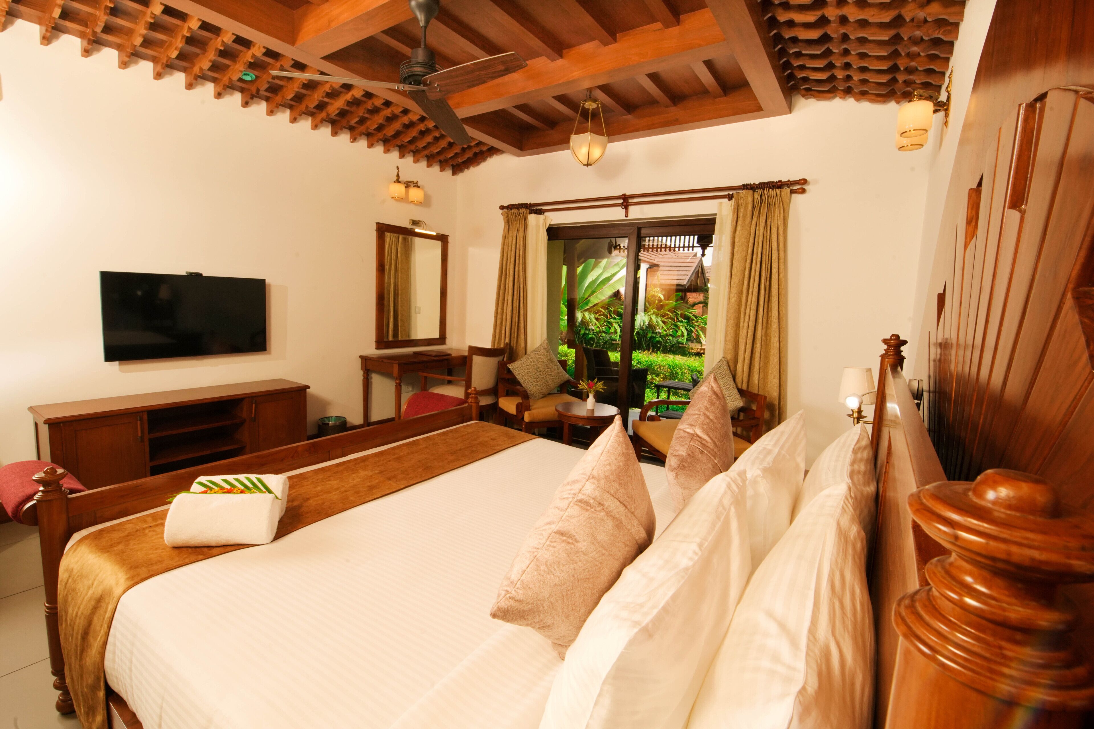 A hotel room with a large bed, wooden headboard, a flat-screen TV, a wooden cabinet, and a view of greenery outside.