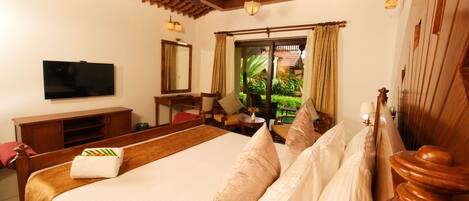 A hotel room with a large bed, wooden headboard, a flat-screen TV, a wooden cabinet, and a view of greenery outside.