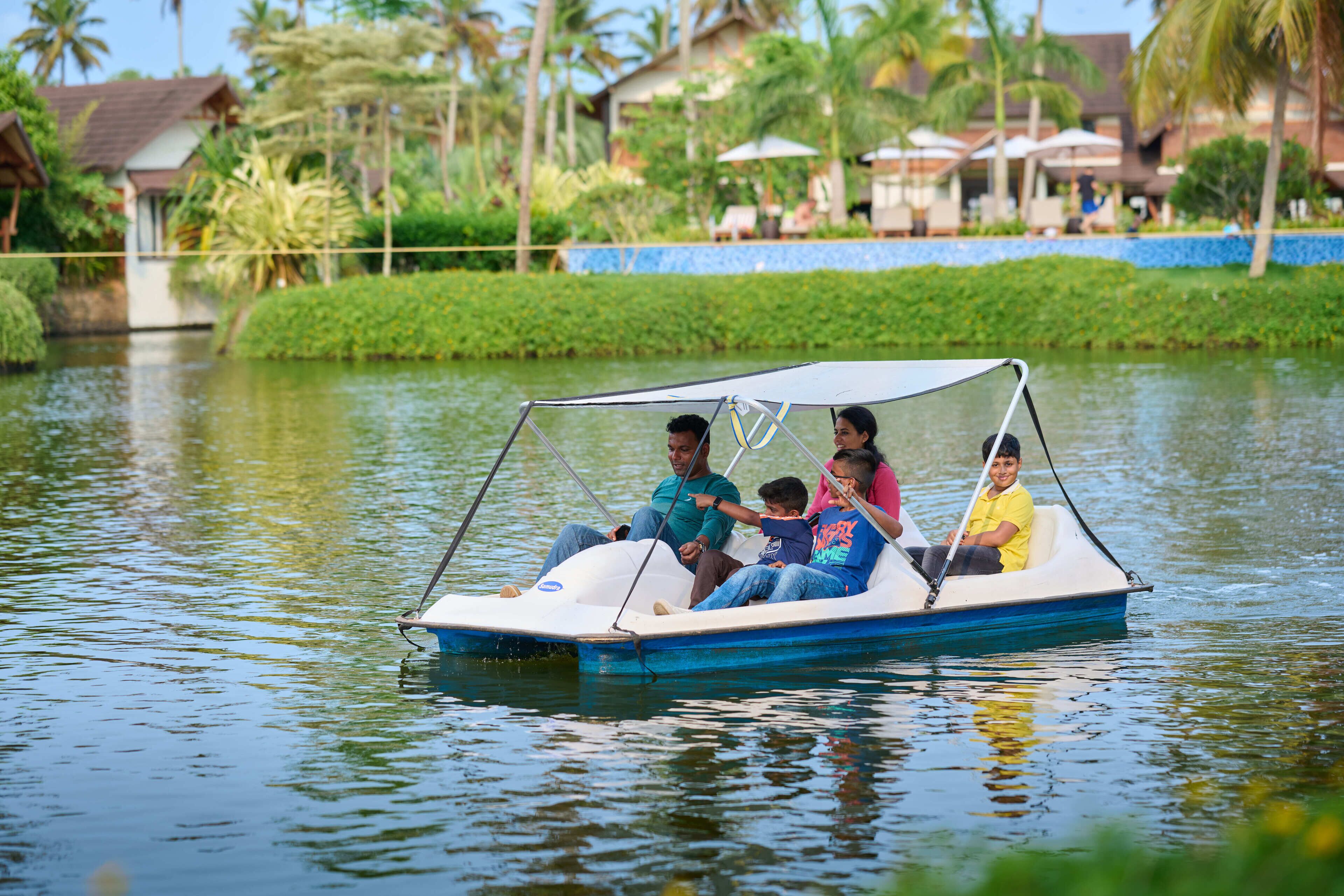 A small boat with four people on a calm water body, surrounded by greenery and buildings in the background.