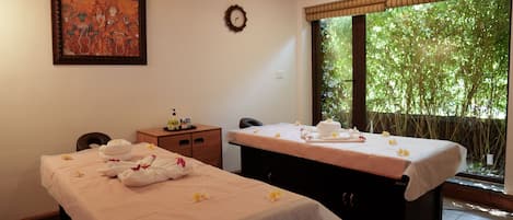 A spa room with two massage tables, a clock, a painting, and a view of greenery through the window.