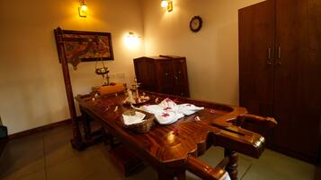 A dining area with a long wooden table set for a meal, flanked by chairs, with a wall-mounted clock and decorative wall sconces.