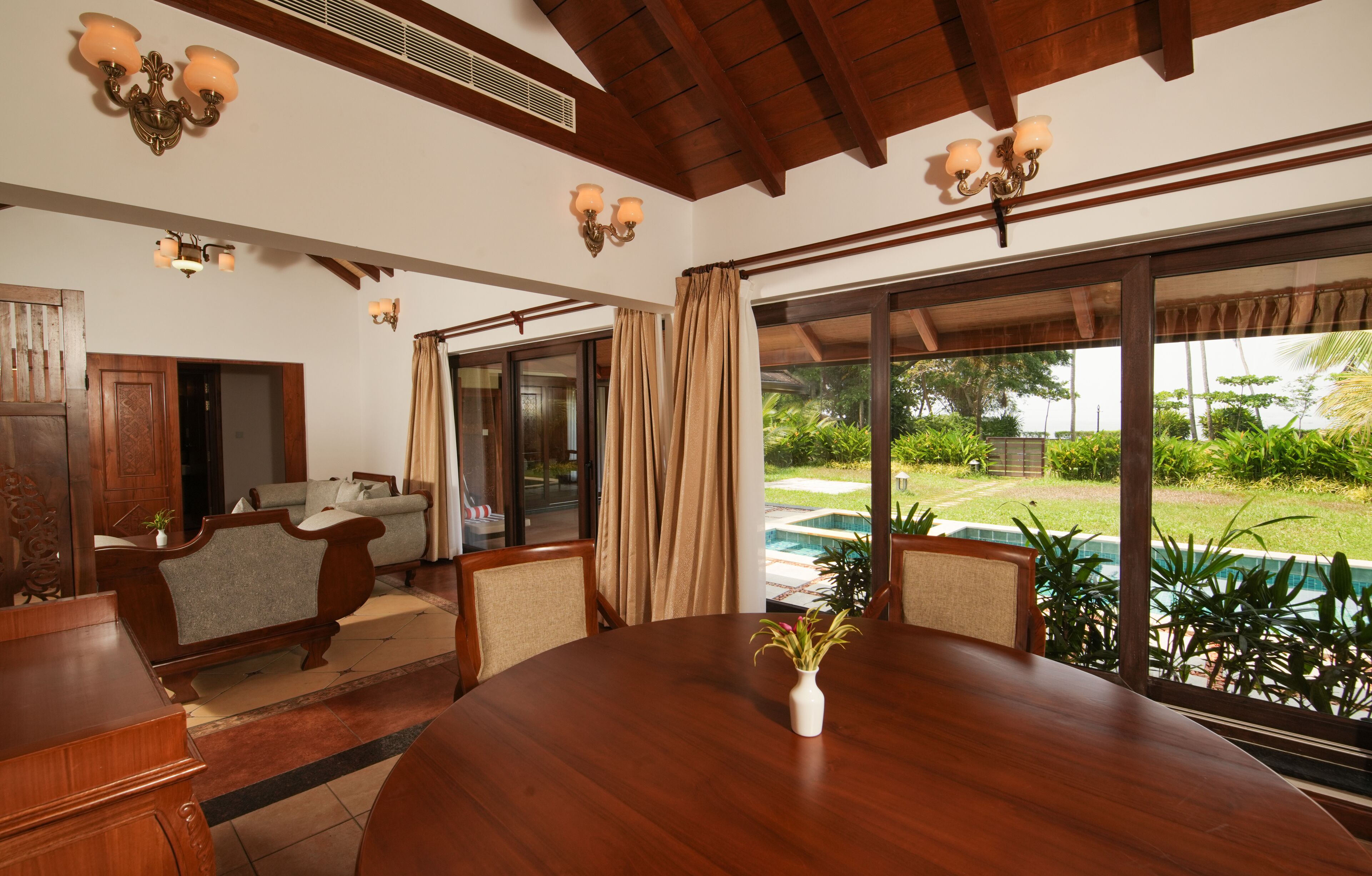 A dining area with a wooden table and chairs, a vase of flowers, and a view of a swimming pool and garden.