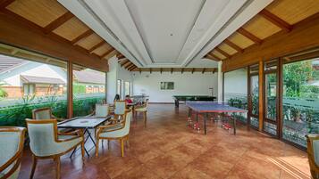 A spacious indoor area with a ping pong table, wooden chairs, and large windows offering views of the outside.