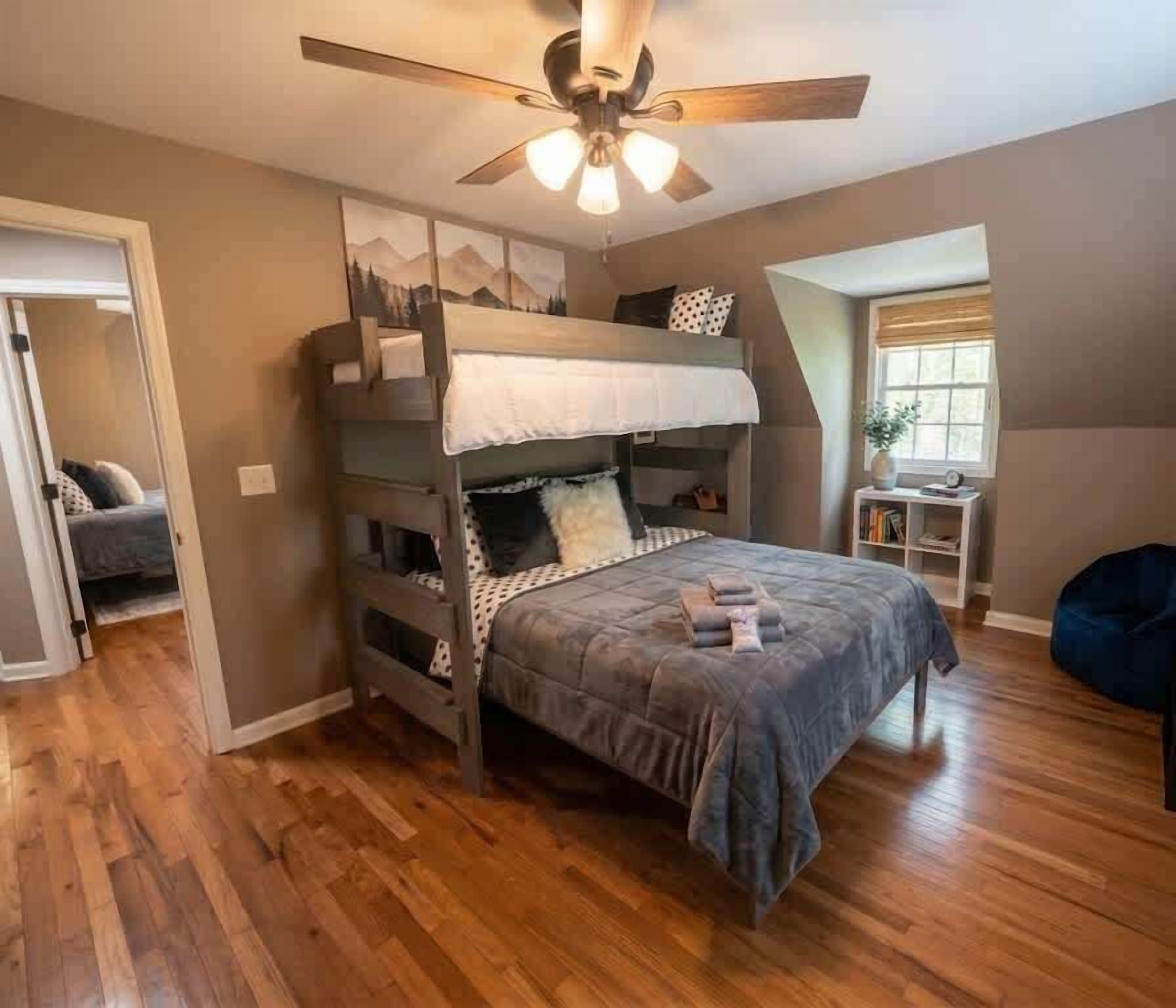 Une chambre avec un lit superposé, un ventilateur de plafond, une petite table avec des livres et une fenêtre avec des rideaux.