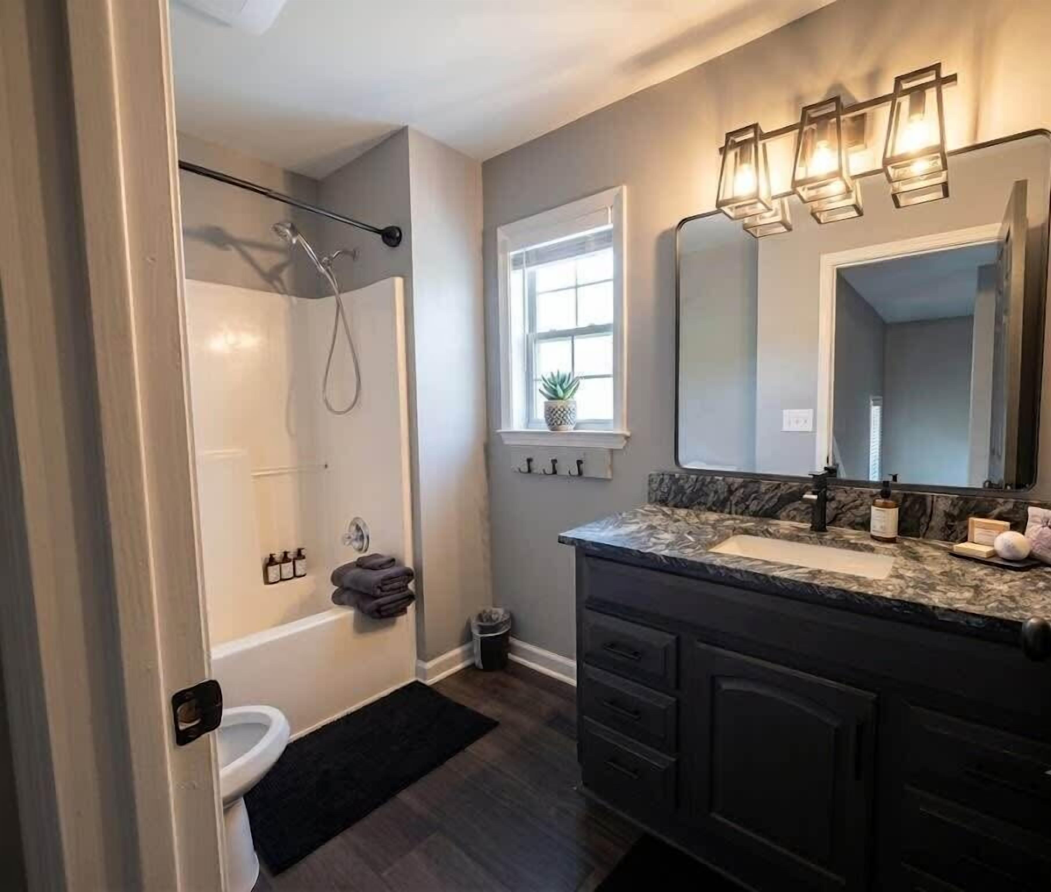 A bathroom with a shower, toilet, vanity with a mirror, and a window.
