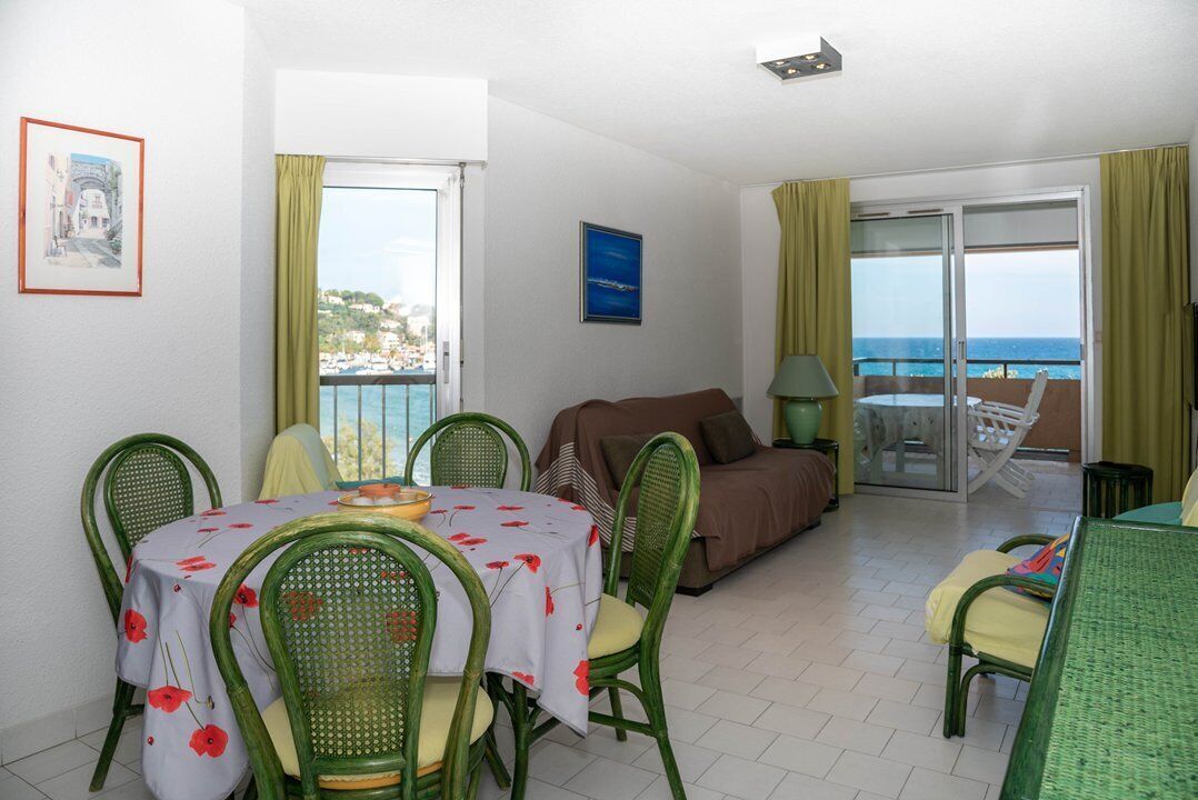 A room with a dining table, chairs, a sofa, and a balcony with a view of the sea.