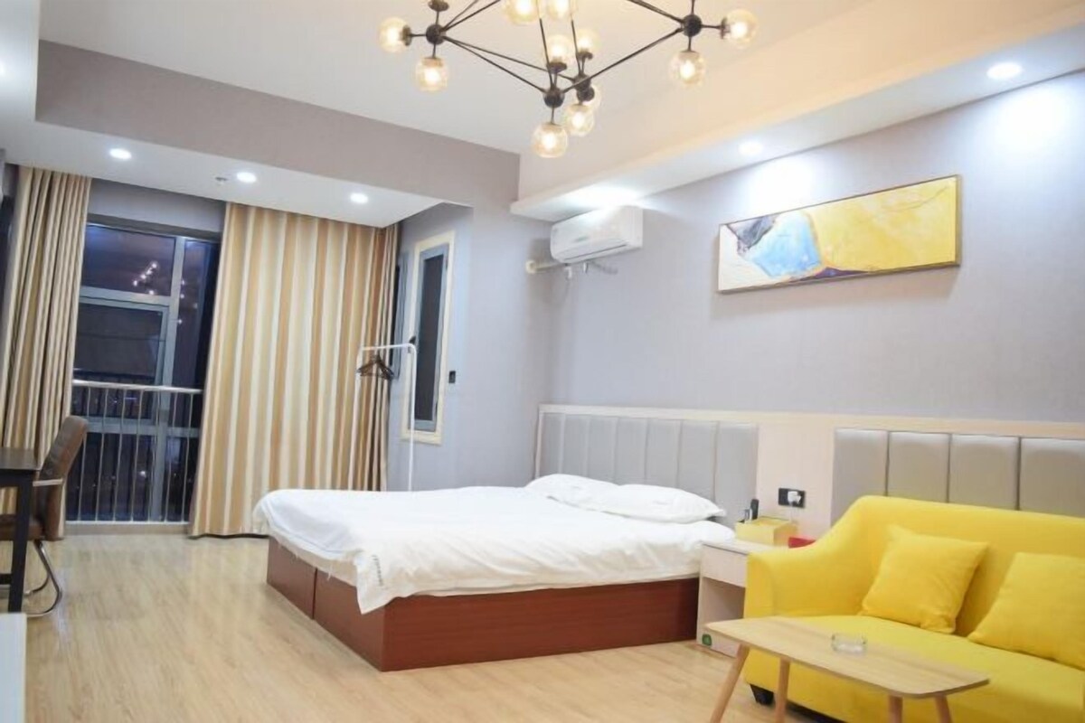 Habitación de hotel con cama, sofá amarillo, una mesita, balcón con cortinas e iluminación en el techo.