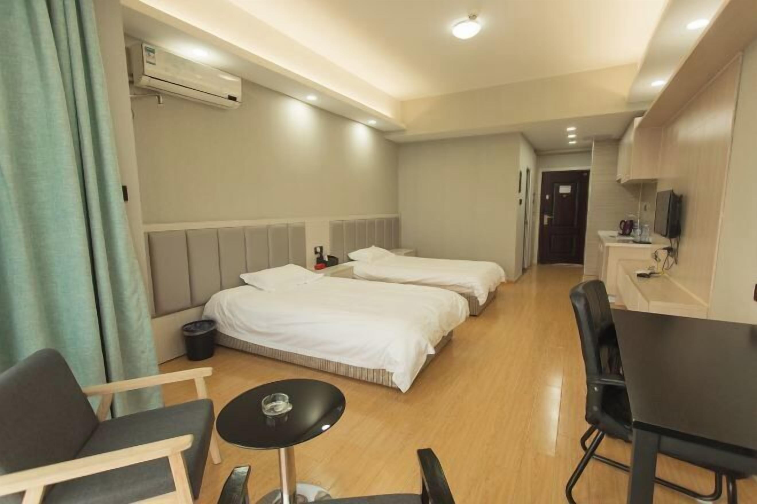 Habitación de hotel con dos camas, una mesita, una silla y un pequeño rincón de cocina.