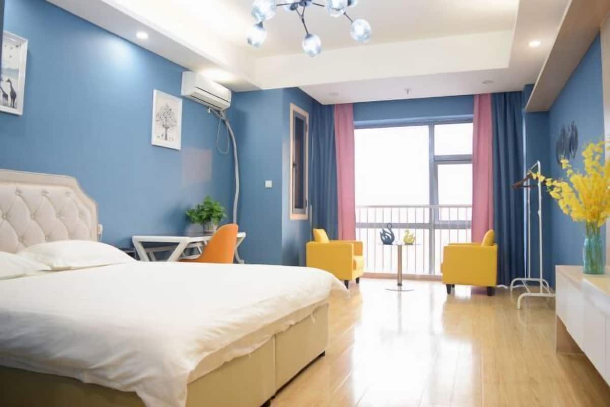 Un dormitorio con una cama, un escritorio, dos sillas, una planta en maceta y un ventanal con cortinas.