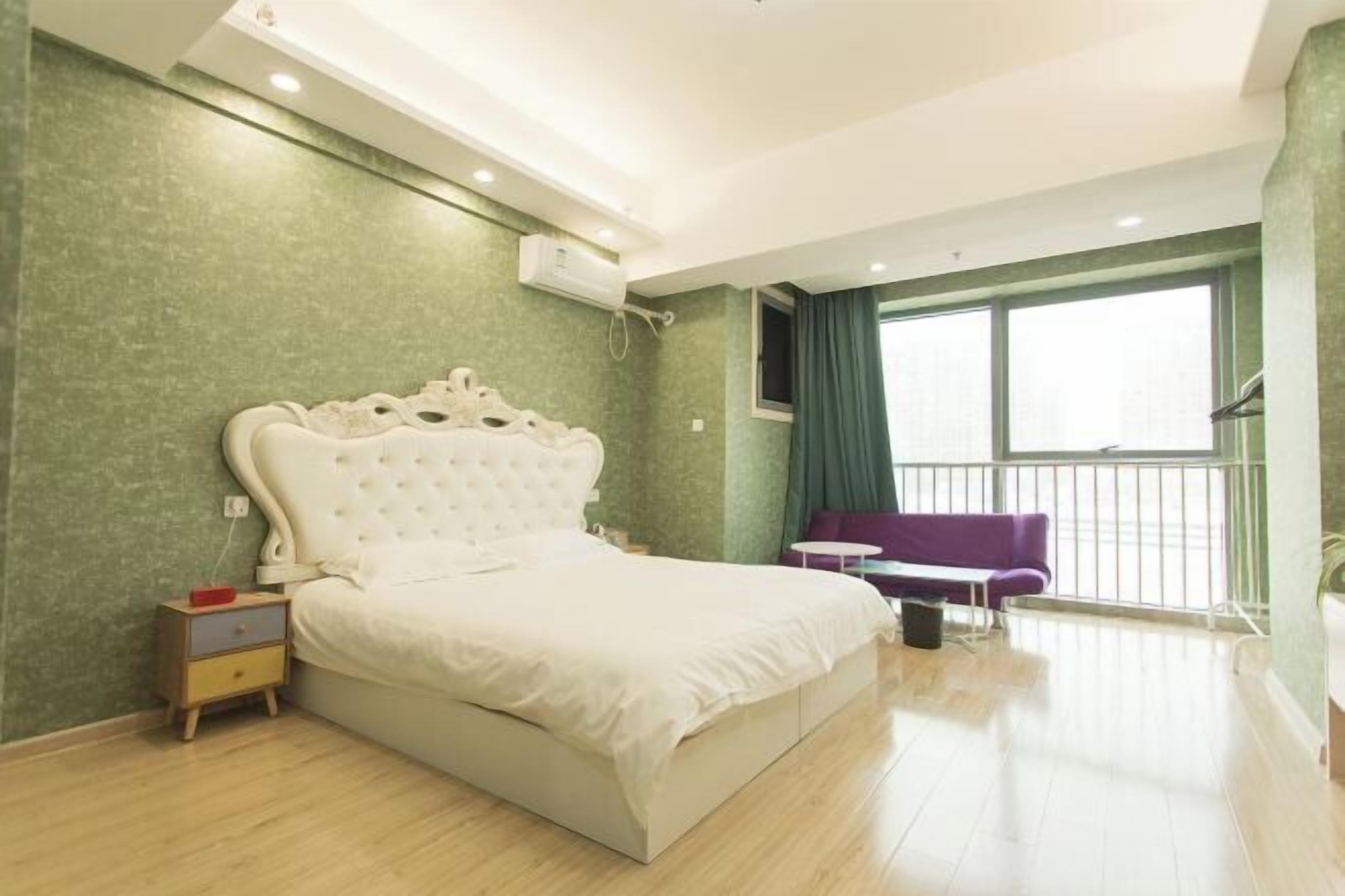 A bedroom with a large bed, a nightstand, a small table, a purple sofa, and a window with a view.