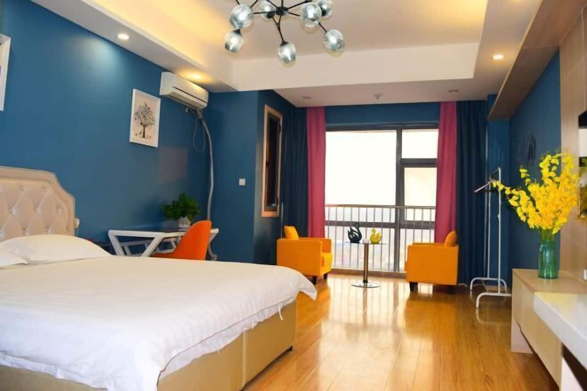 Un dormitorio con cama, escritorio, dos sillas, balcón con barandilla y un florero con flores.