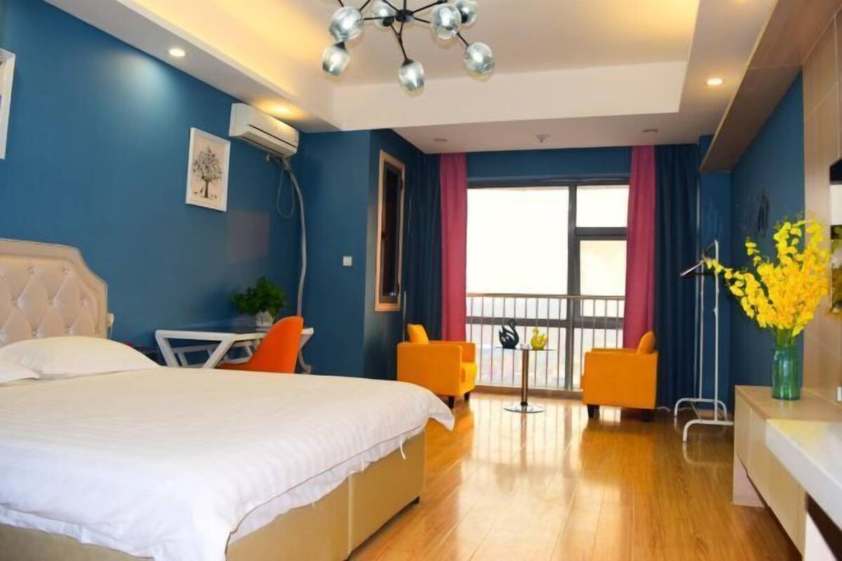 A bedroom with a bed, a desk, two chairs, a balcony with a railing, and a vase with flowers.