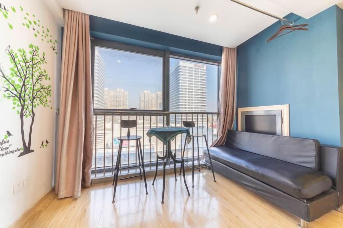 A modern living room with a black leather sofa, a small table, and two chairs. A large window offers a view of tall buildings.