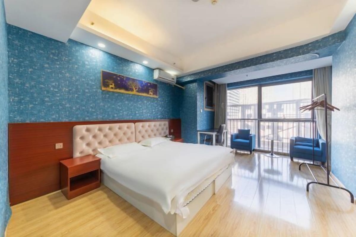 A hotel room with a large bed, a wooden nightstand, a desk with a chair, and a balcony with a view.