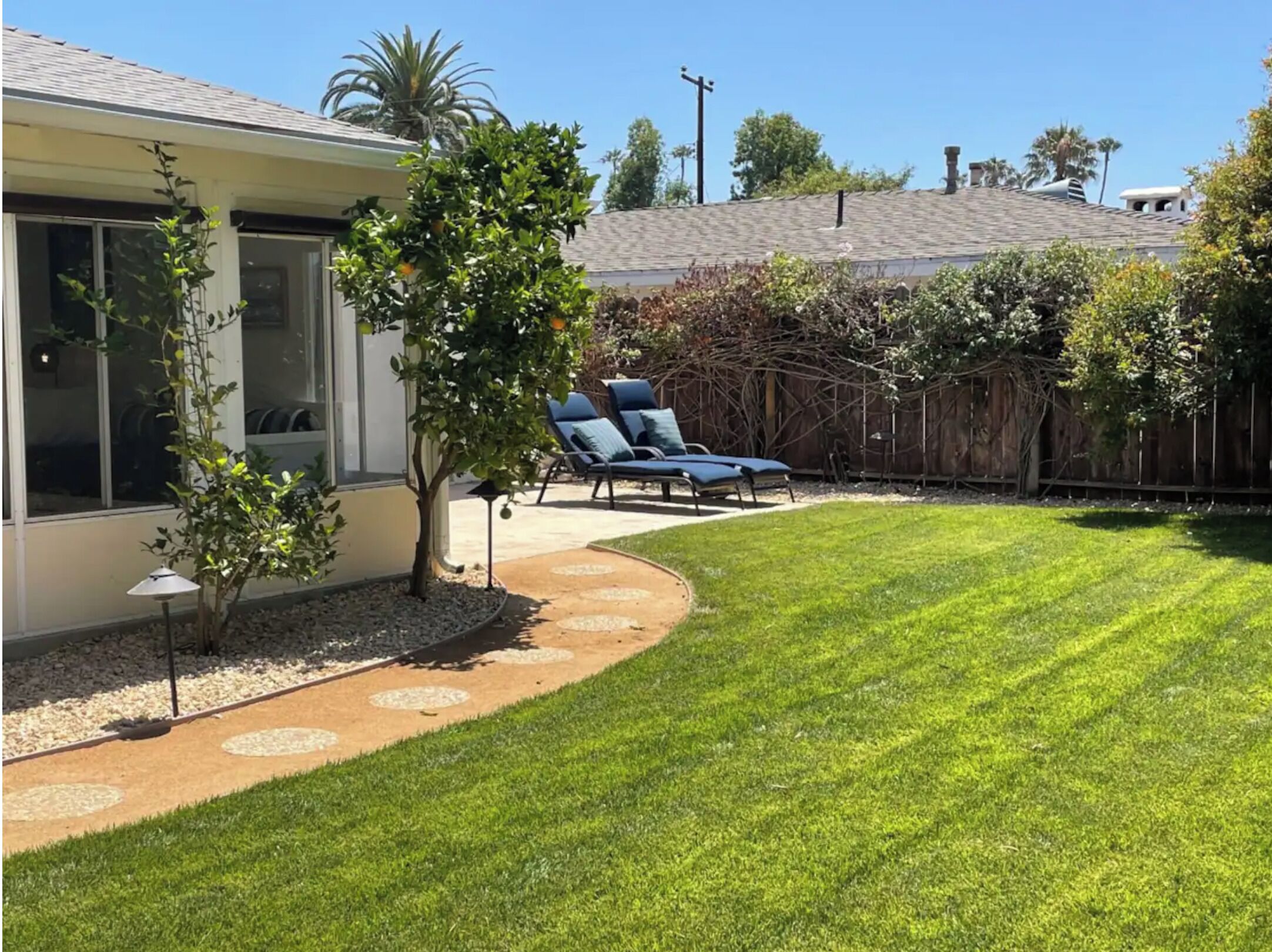 A backyard with a lawn, a tree, a patio with chairs, and a fence.