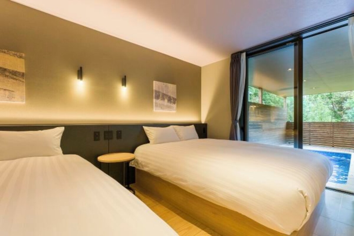 A modern hotel room with two beds, a small round table, and a large window offering a view of the outdoors.