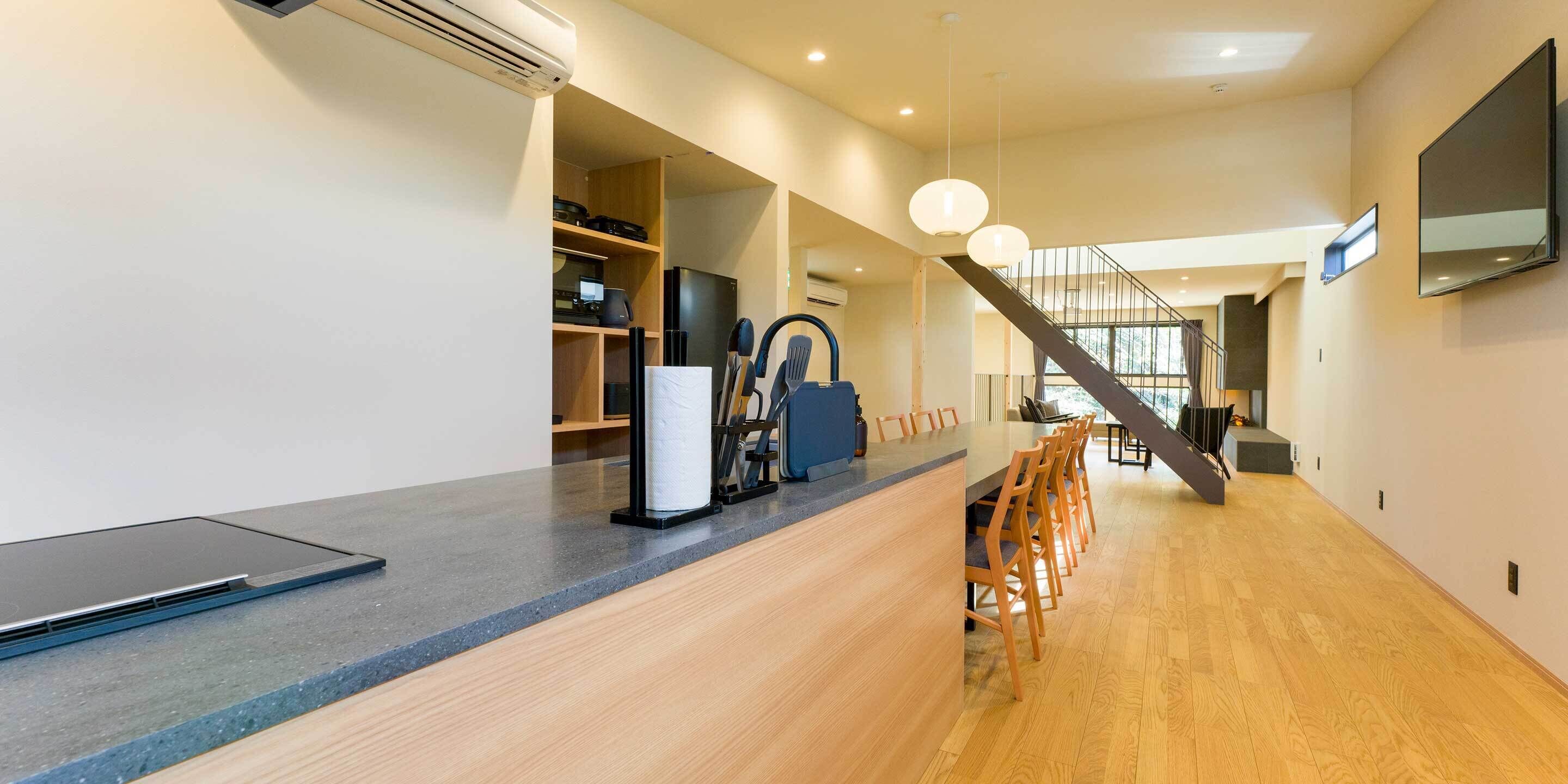 A modern kitchen with a built-in laptop, a coffee machine, and a refrigerator. There is a wooden dining area with chairs and a staircase leading to another level.