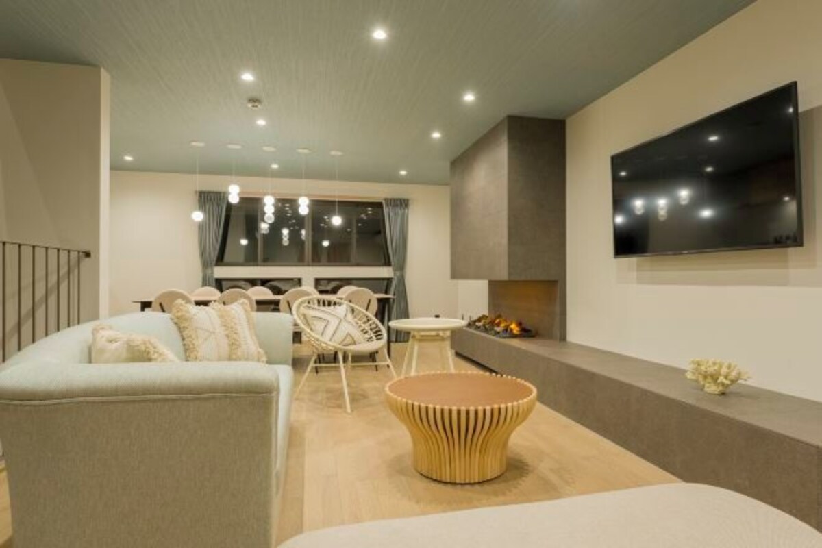 A modern living room with a sofa, chairs, a coffee table, a flat-screen TV mounted on the wall, and a fireplace.