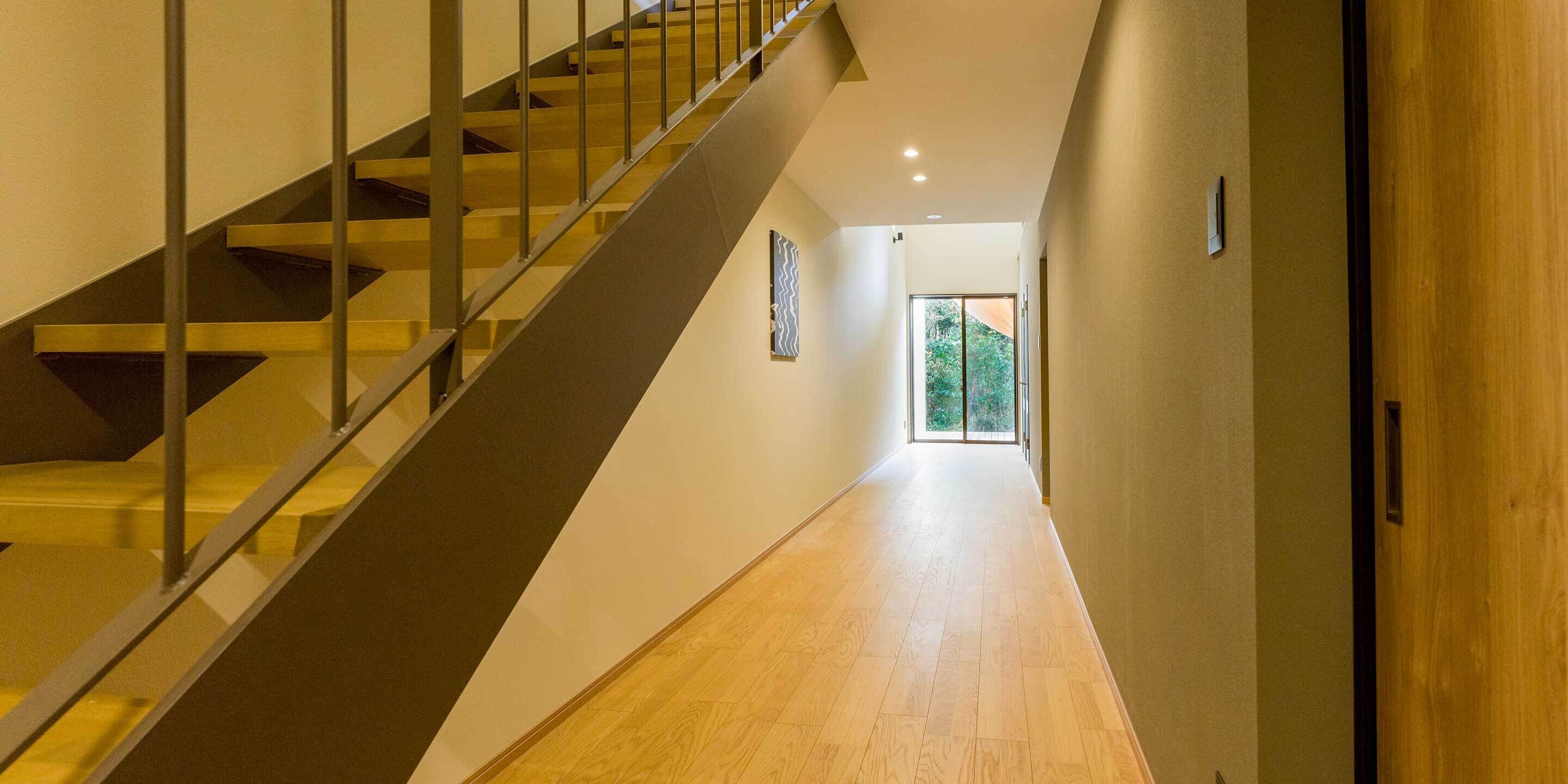 A modern interior with a wooden staircase, a hallway leading to a door, and a view of greenery outside.
