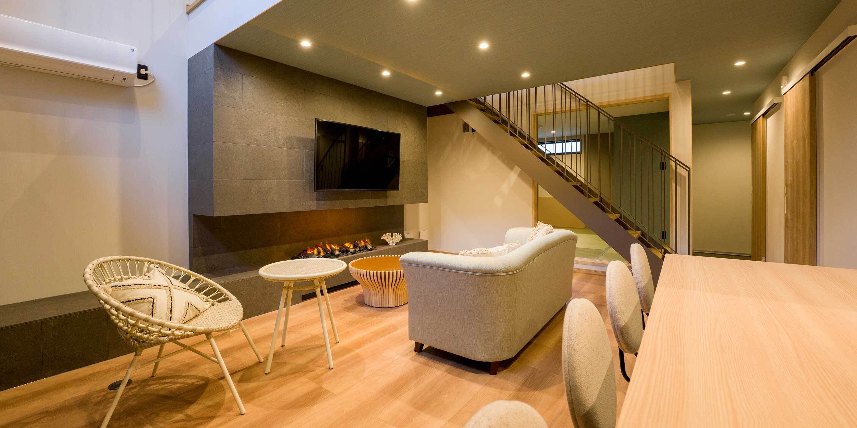 A modern living room with a sofa, a round table, a wicker chair, a flat-screen TV mounted on the wall, and a staircase leading to another level.