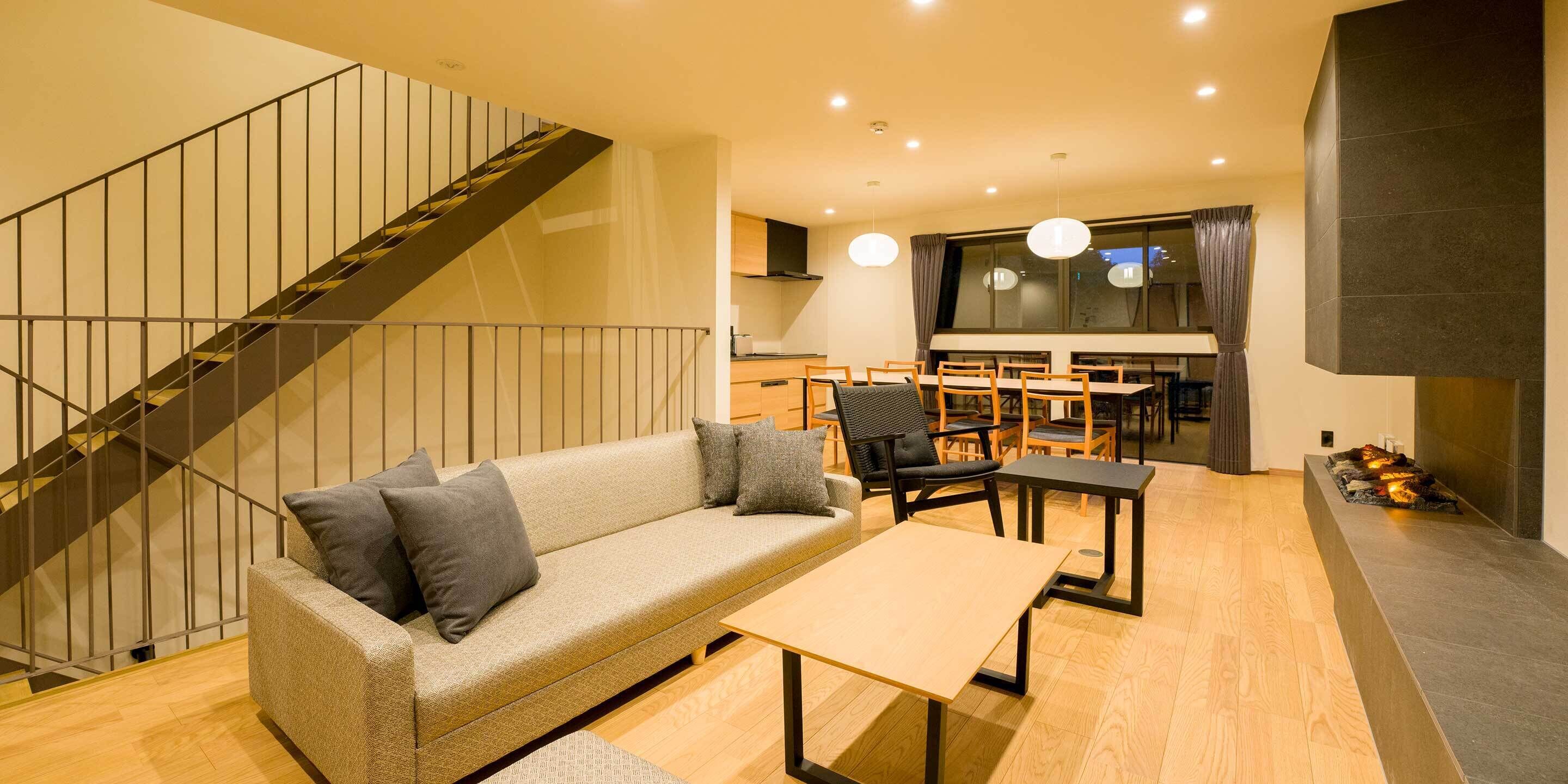 A modern living room with a sofa, a coffee table, and a dining area with chairs and a table. A staircase with a railing is visible in the background.