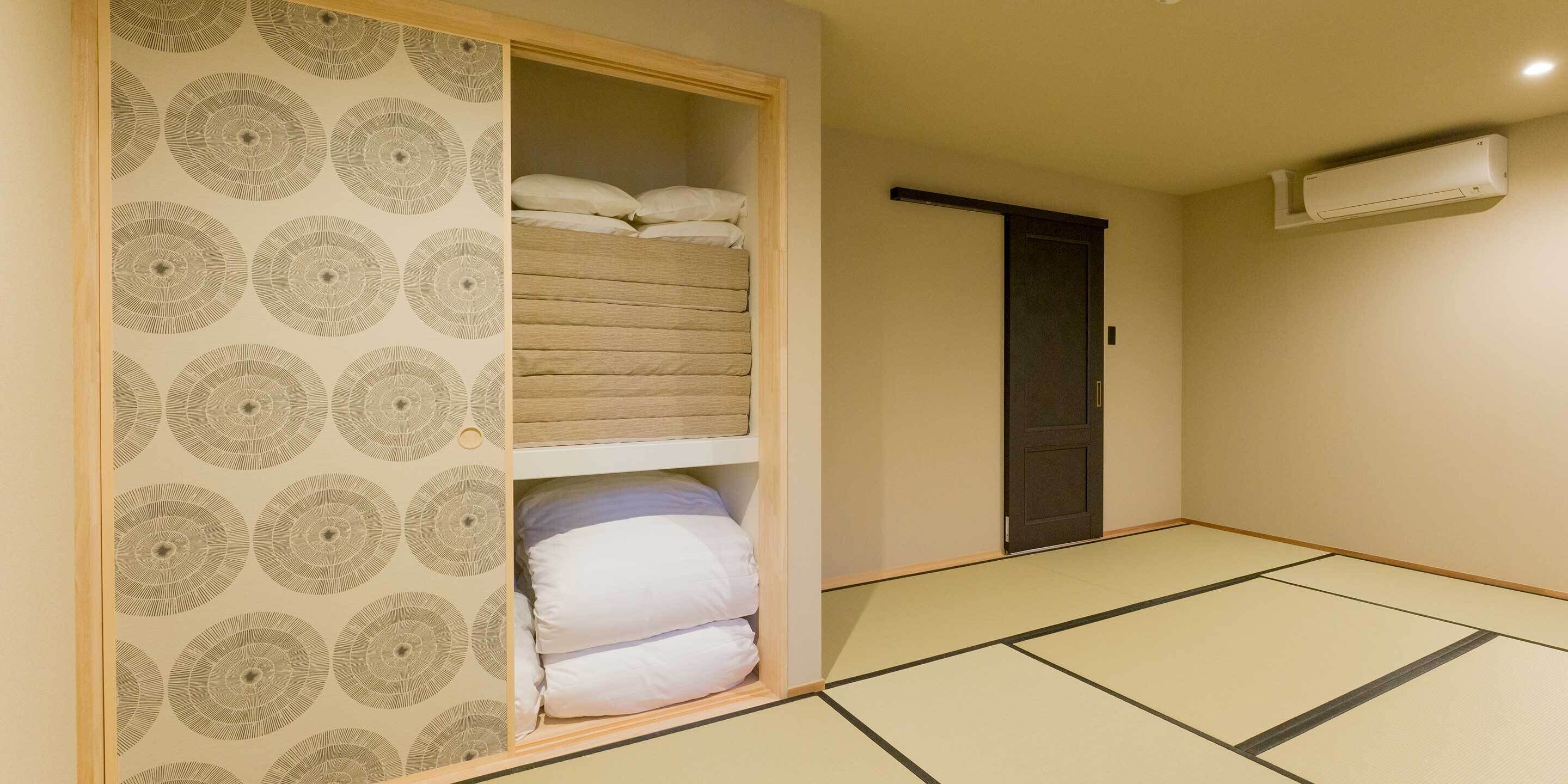 A room with tatami flooring, a sliding door with patterned wallpaper, a bed with white bedding, and an air conditioning unit on the wall.