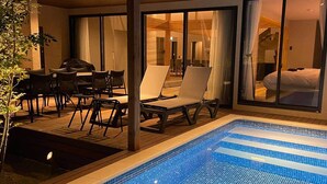 Uma área de piscina com espreguiçadeiras e uma mesa de jantar, grandes portas de vidro deslizantes que dão para um quarto e uma escada visível.