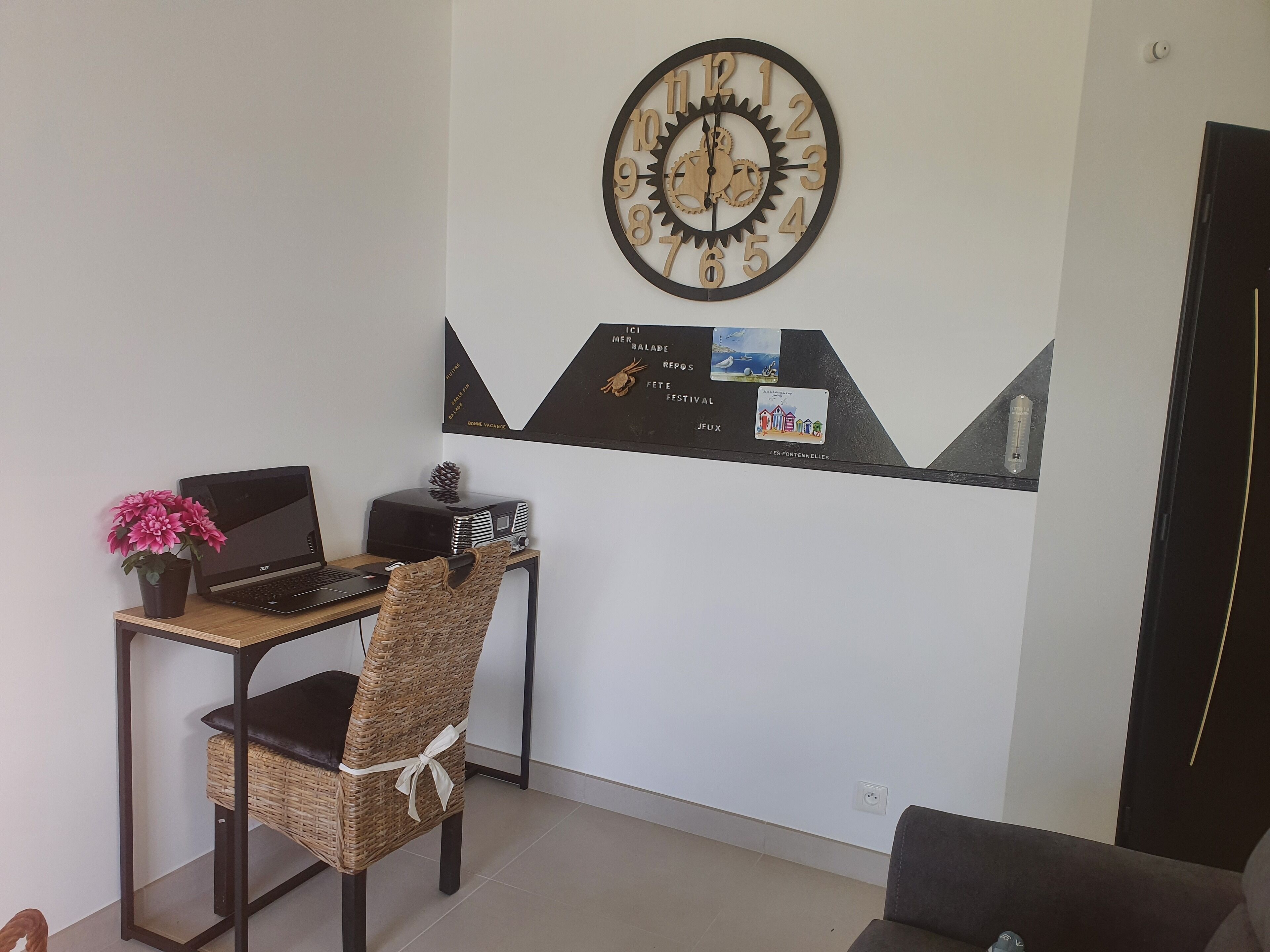 Una sala de estar moderna con un ordenador portátil sobre un escritorio, una silla de mimbre, un reloj de pared decorativo y una obra de arte colgada en la pared.