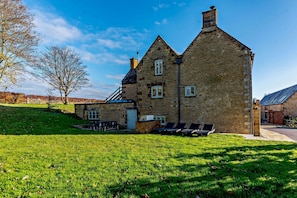 A stone building with a steep roof, a small attached structure, and a grassy lawn with outdoor seating. - 8 bedroom luxury holiday home in the Cotswolds with a hot tub - Stonewell Farmhouse (Great Tew)